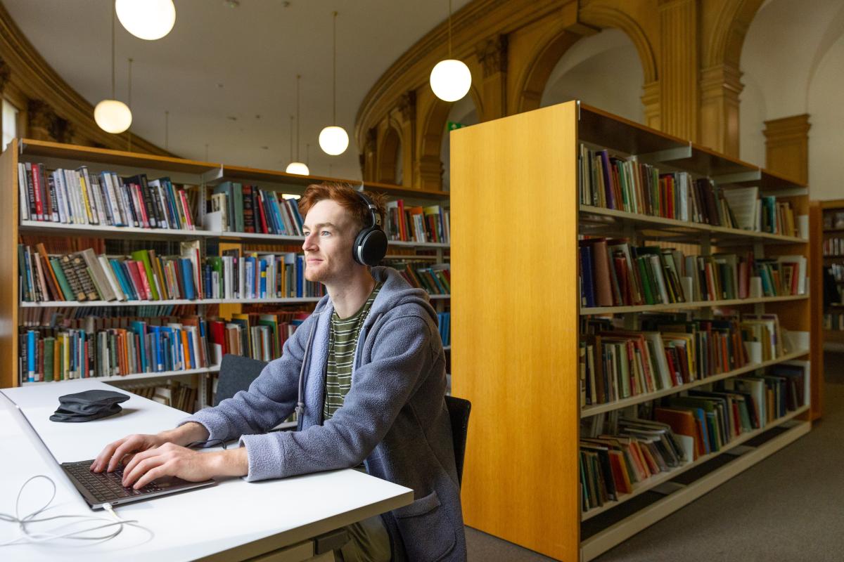 Young man with headphones on using a laptop with library bookshelves behind