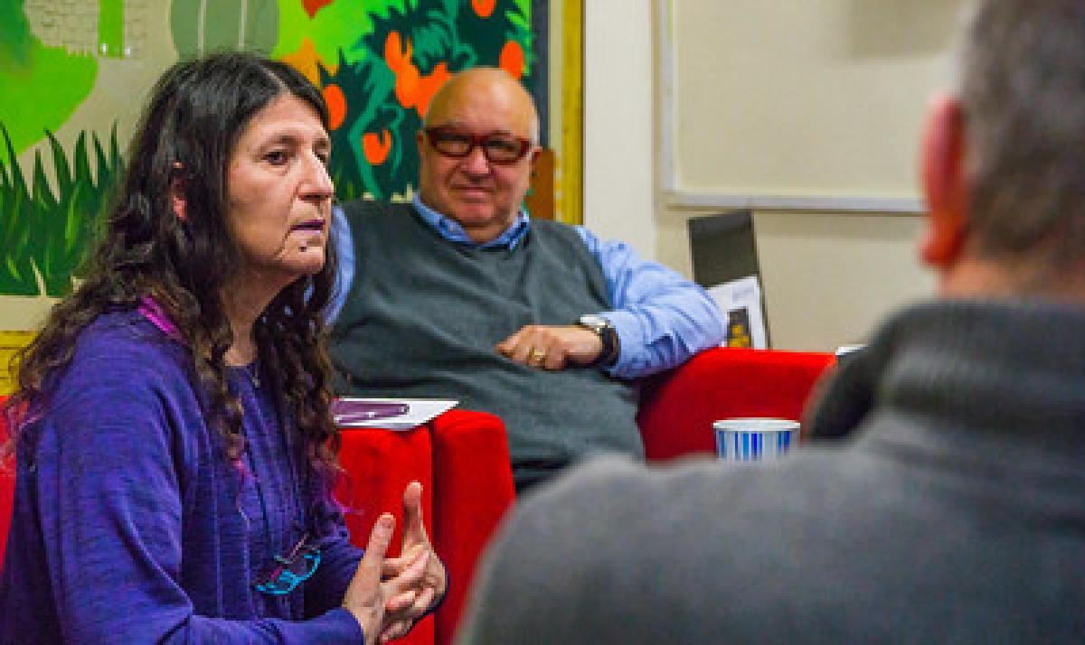 A man in a grey sleeveless sweater listens to a woman speaking.