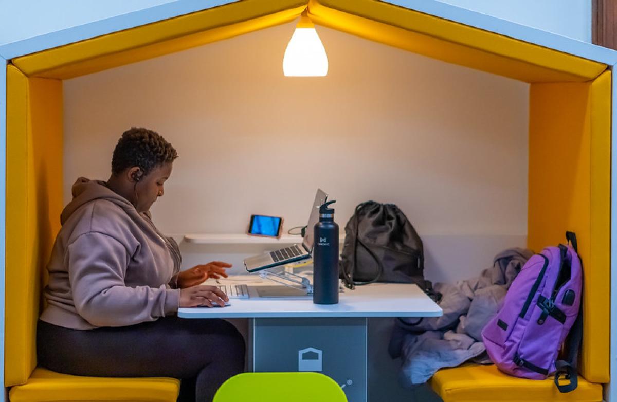 A young girl sits studying at a laptop in a library cubby hole. Above her a lamp shines.