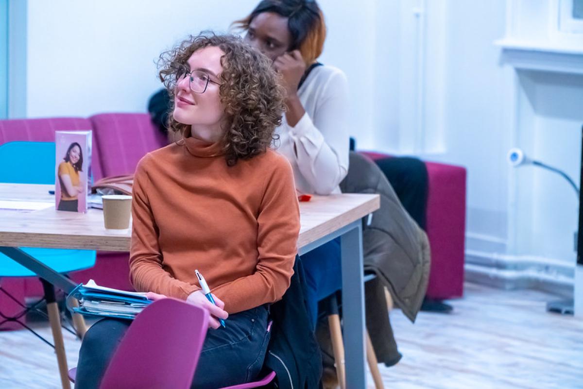 A woman dressed in a terracotta coloured sweatshirt smiles slightly as she listens to a presentation.