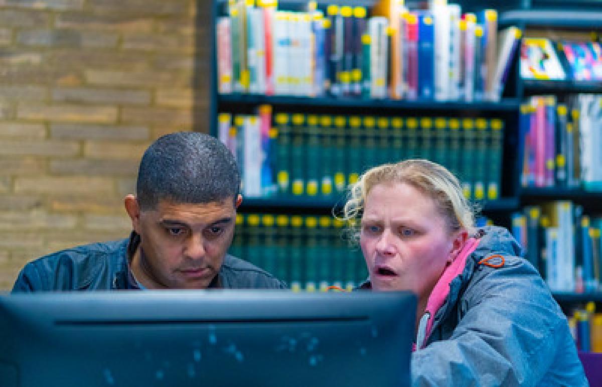 A woman wearing a coat with a pink lining helps a man use a computer. Behind them is a large bookshelf.