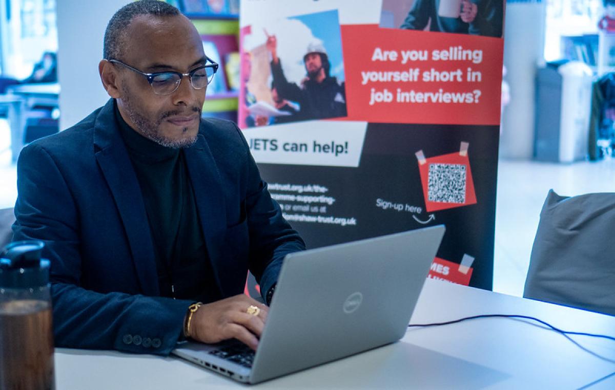 A man wearing a gold ring on his middle finger works at a laptop in a library