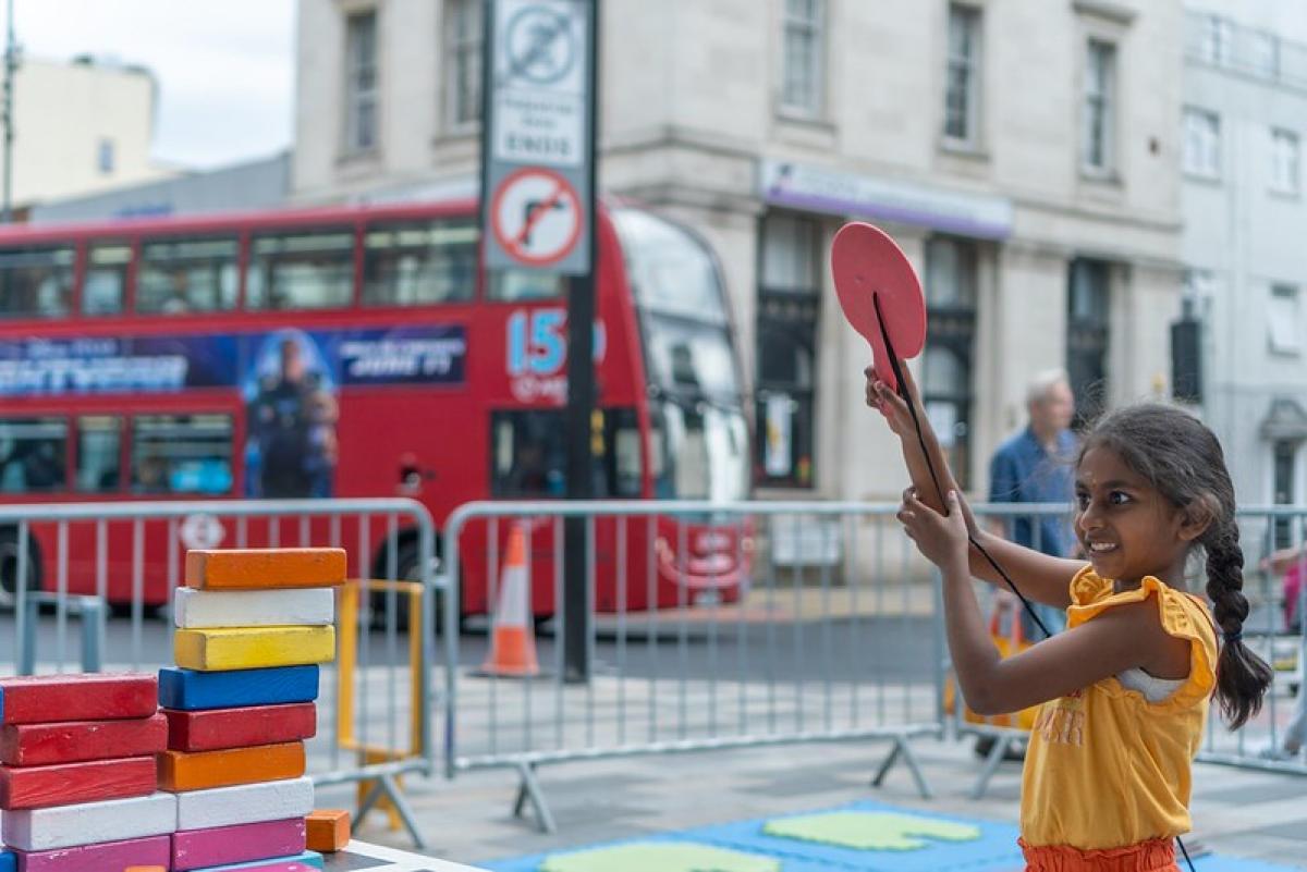 A young girl holds up a red bat with a smile on her face. In the background is a red London bus.