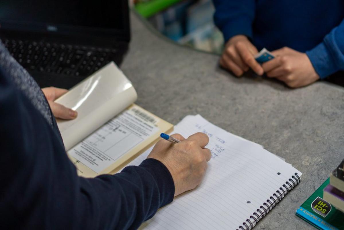 A hand writes on a pad of paper. A library book is open at the issue page to the left of the pad.