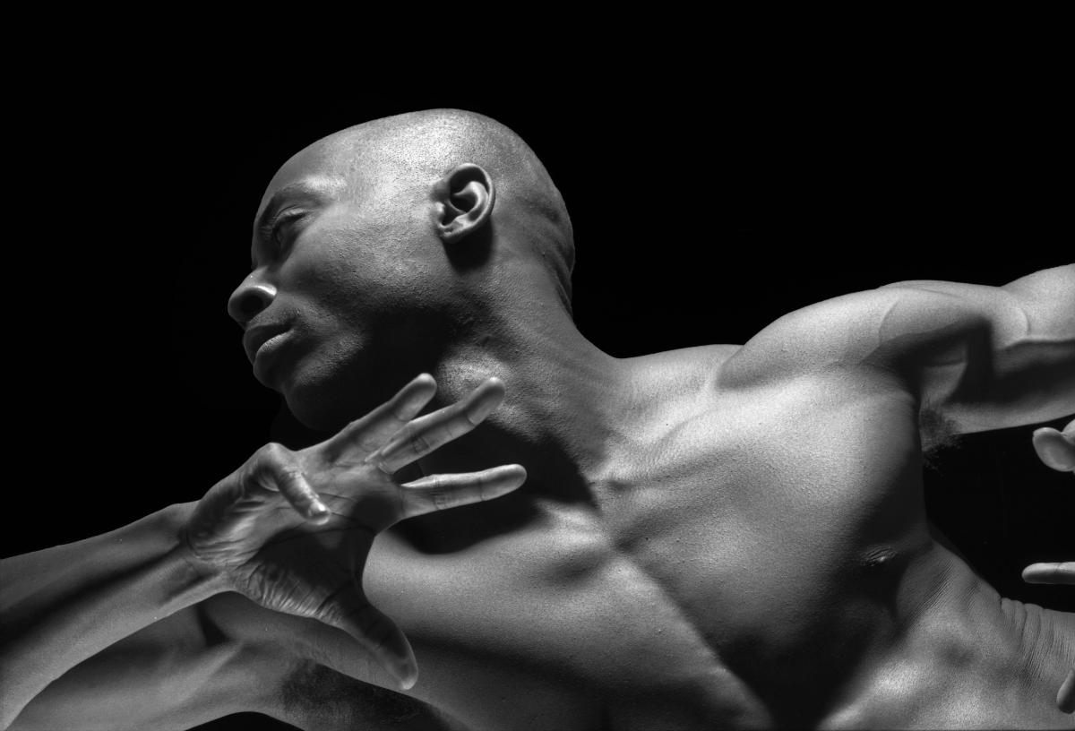 This black-and-white image shows a muscular man in a dynamic pose, with his head turned and arm raised. The lighting highlights his strong features and defined muscles against a dark background.