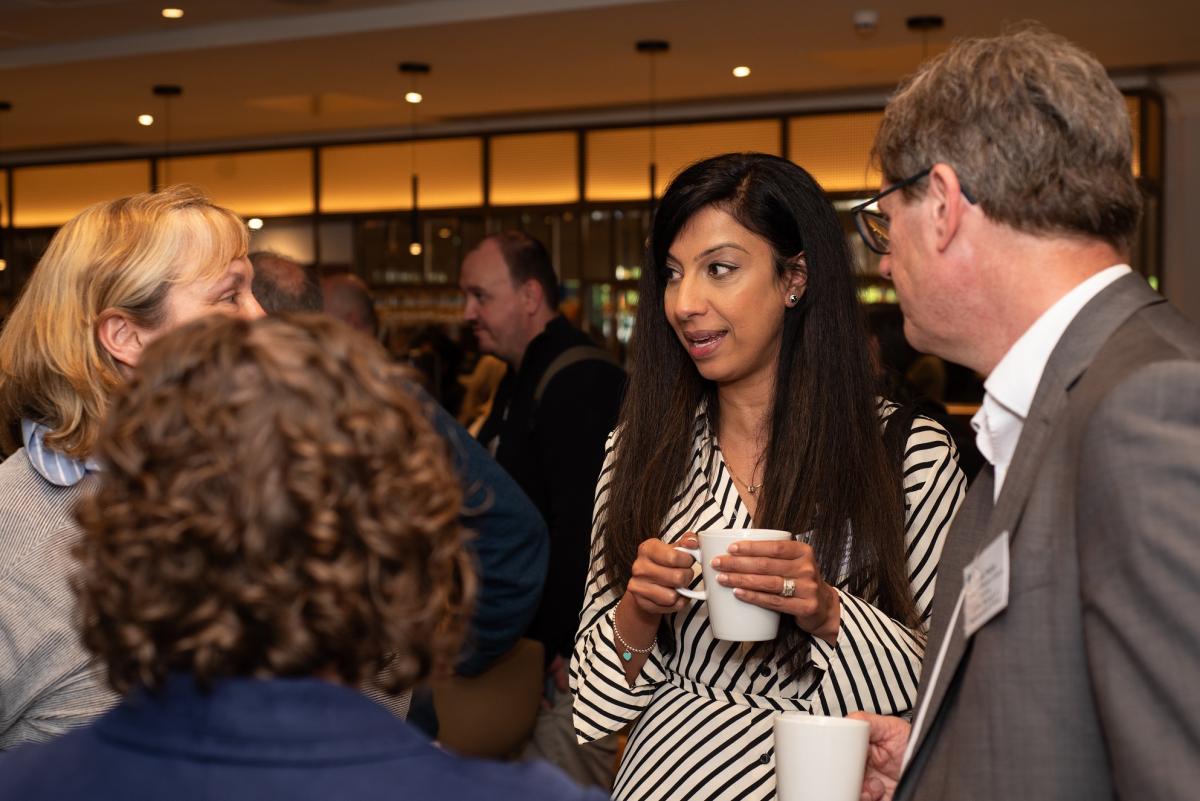 The image shows a group of people engaged in conversation during the Libraries Connected Annual Seminar. A woman in a black-and-white striped blouse is holding a white mug and actively listening or speaking to two individuals near her. The background is blurred, with other participants visible in the setting, giving a warm and casual atmosphere.