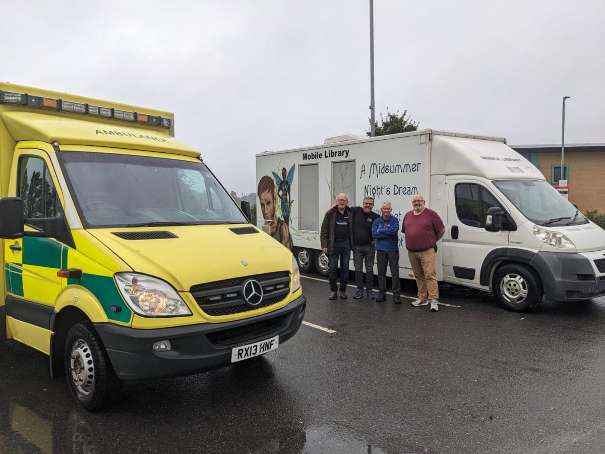 An ambulance stands in front of a mobile library. In front of the library are three men.