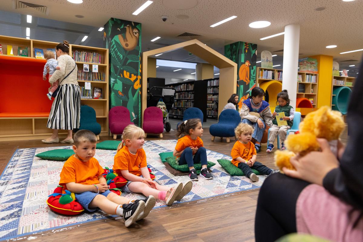 Several children dressed in orange listen to a storyteller in a library