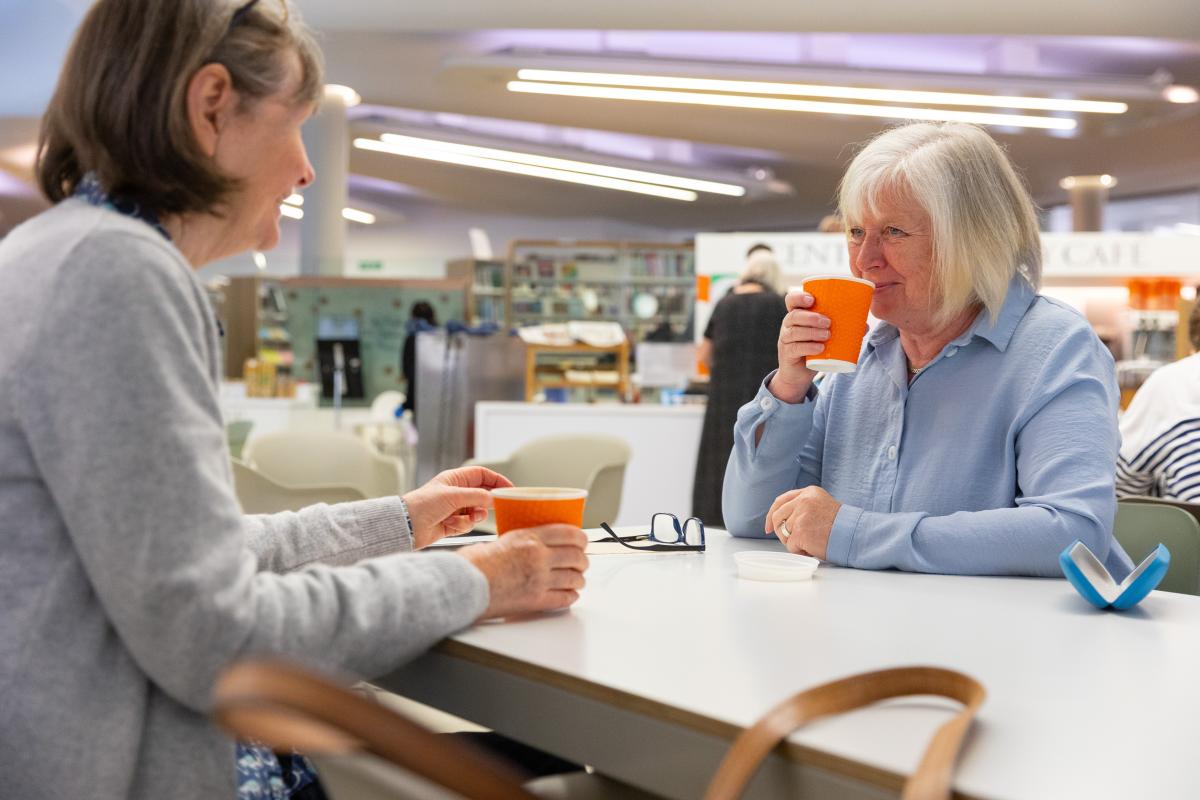 A woman speaks to a female librarian with a cup of hot drink in her hand.