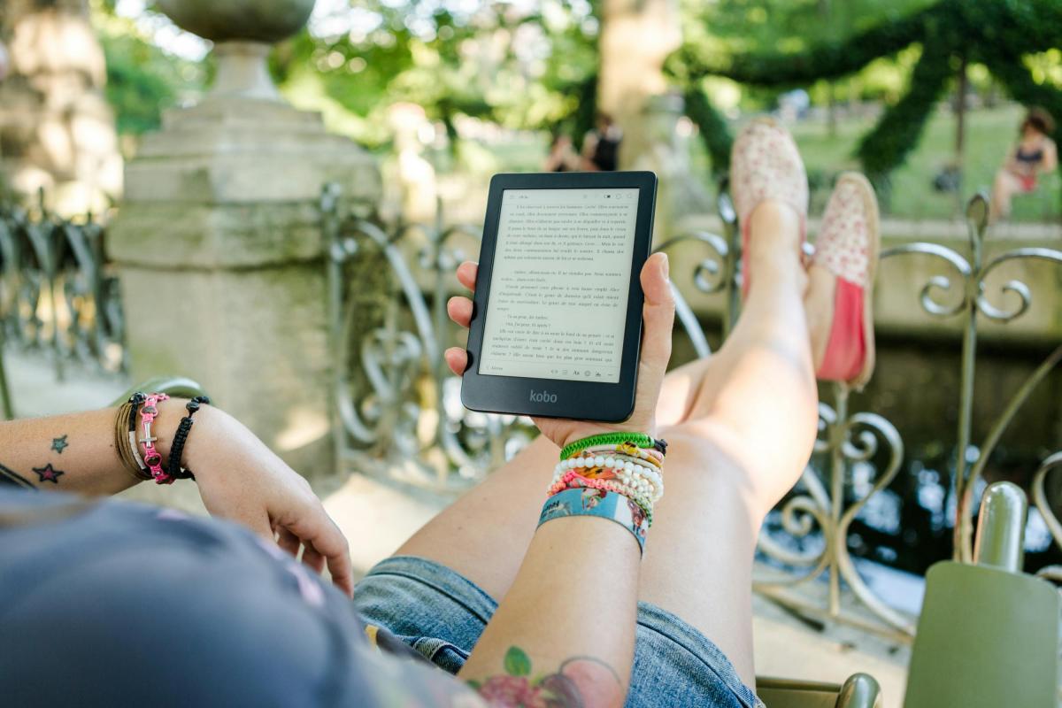 A person is relaxing outdoors, reclining on a chair with legs stretched out and crossed at the ankles, wearing denim shorts and pink floral slip-on shoes. They are holding an ereader in one hand, displaying text in French. The person has colorful bracelets and wristbands on both wrists, along with star tattoos. In the background, there is an ornate metal railing, stone structures, and greenery, suggesting a park or garden setting with blurred figures in the distance.