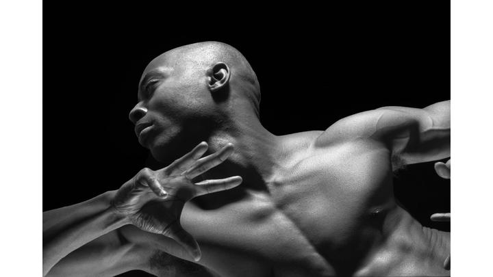 This black-and-white image shows a muscular man in a dynamic pose, with his head turned and arm raised. The lighting highlights his strong features and defined muscles against a dark background.