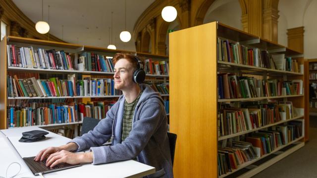 Young man with headphones on using a laptop with library bookshelves behind