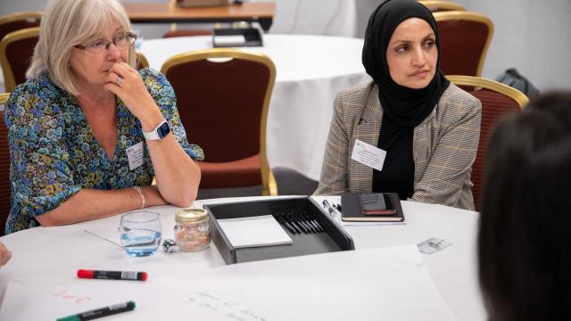 A group of two women engaged in thoughtful discussion during a meeting, with writing tools and notes on the table in front of them.