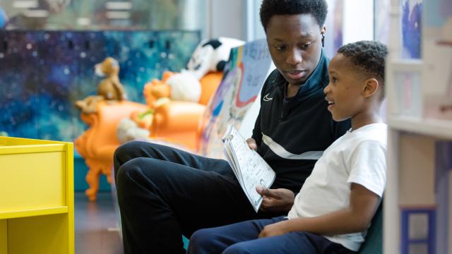 A man is sitting with a young boy, reading together in a bright, colourful children's library.