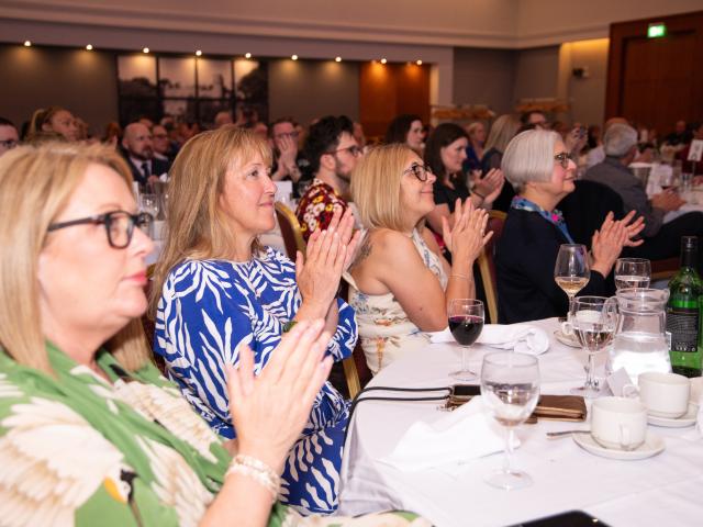 Four women are seated around a table at an awards ceremony. They are looking towards the stage and clapping.