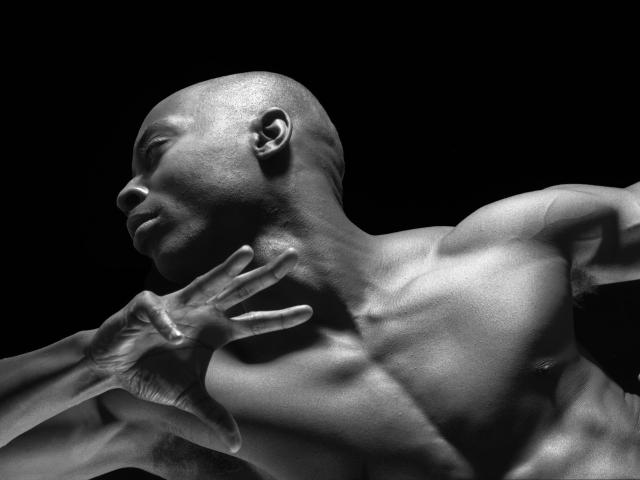 This black-and-white image shows a muscular man in a dynamic pose, with his head turned and arm raised. The lighting highlights his strong features and defined muscles against a dark background.