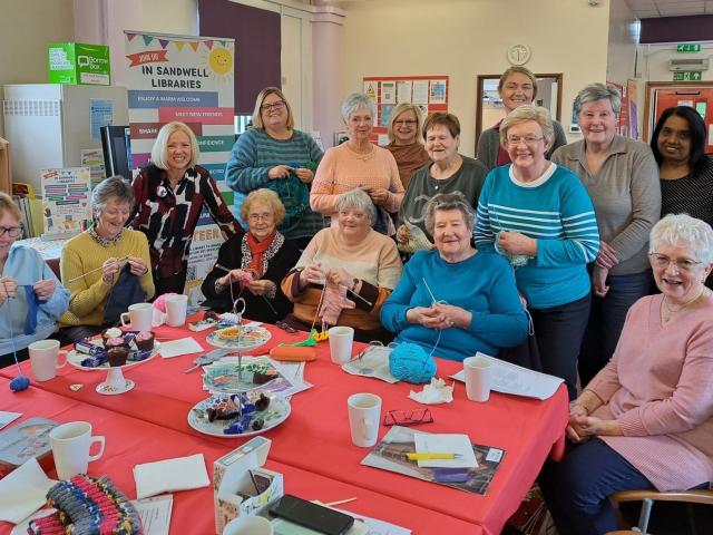 A group of people gathered around a table in a library, enjoying a knitting and social gathering.