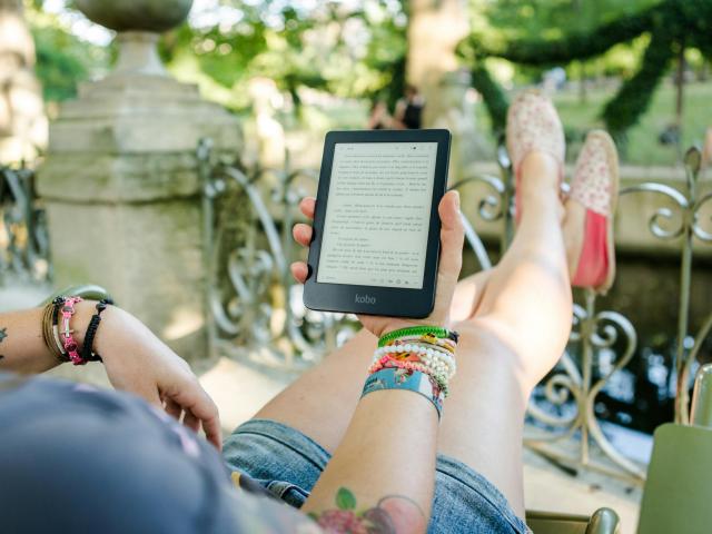 A person is relaxing outdoors, reclining on a chair with legs stretched out and crossed at the ankles, wearing denim shorts and pink floral slip-on shoes. They are holding an ereader in one hand, displaying text in French. The person has colorful bracelets and wristbands on both wrists, along with star tattoos. In the background, there is an ornate metal railing, stone structures, and greenery, suggesting a park or garden setting with blurred figures in the distance.