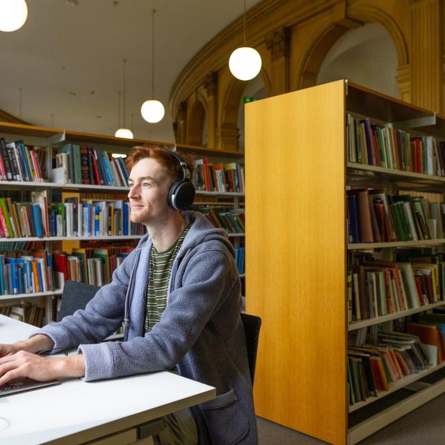 Young man with headphones on using a laptop with library bookshelves behind