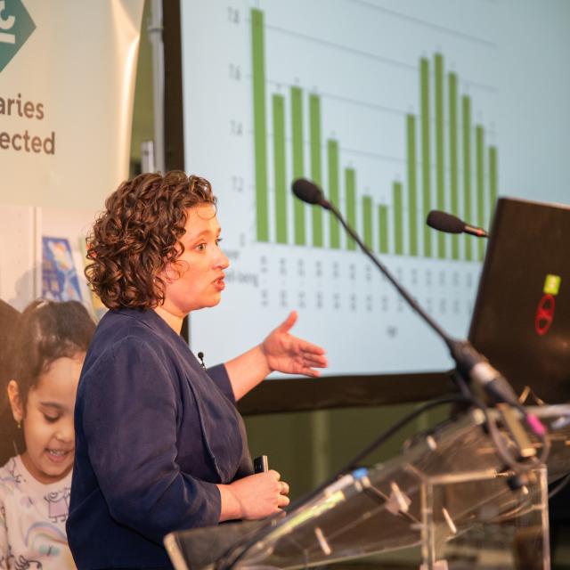 A woman is stood at a lectern giving a presentation wit a bar chart projected on to a screen behind her.