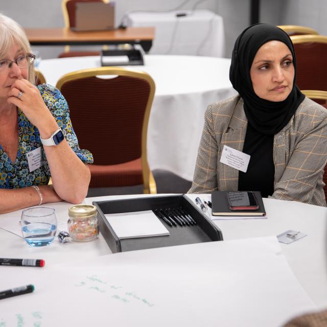 A group of two women engaged in thoughtful discussion during a meeting, with writing tools and notes on the table in front of them.