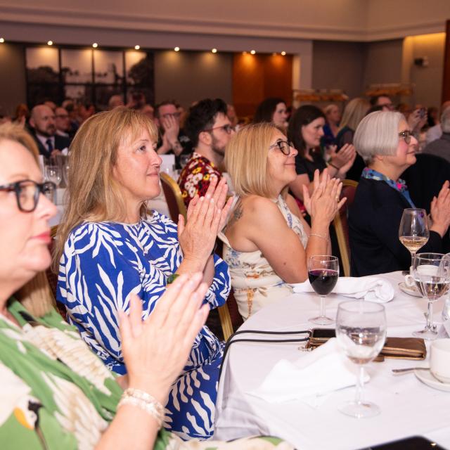 Four women are seated around a table at an awards ceremony. They are looking towards the stage and clapping.