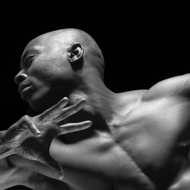 This black-and-white image shows a muscular man in a dynamic pose, with his head turned and arm raised. The lighting highlights his strong features and defined muscles against a dark background.