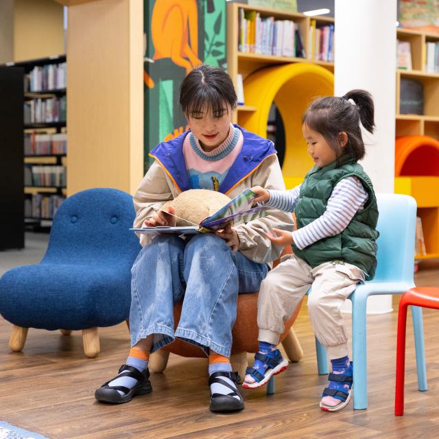 A woman and a young child sit together in a brightly coloured library, sharing a moment as they read a book, with the child excitedly pointing at the pages.