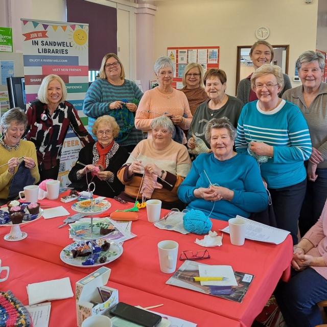 A group of people gathered around a table in a library, enjoying a knitting and social gathering.