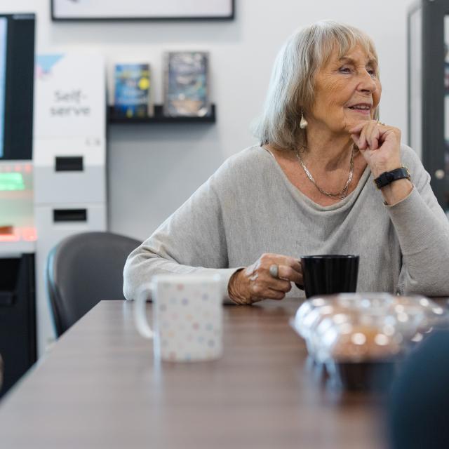 An older woman with light skin and shoulder-length gray hair sits at a table in a modern library space