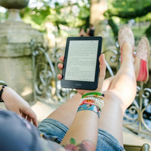 A person is relaxing outdoors, reclining on a chair with legs stretched out and crossed at the ankles, wearing denim shorts and pink floral slip-on shoes. They are holding an ereader in one hand, displaying text in French. The person has colorful bracelets and wristbands on both wrists, along with star tattoos. In the background, there is an ornate metal railing, stone structures, and greenery, suggesting a park or garden setting with blurred figures in the distance.