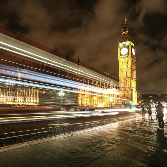 This image captures a nighttime view of Big Ben and the Houses of Parliament in London. The long-exposure photograph shows light trails from passing vehicles on the bridge, creating streaks of white, yellow, and red lights. The illuminated clock tower and buildings contrast against the dark, cloudy sky. Pedestrians are visible on the right side, some appearing blurred due to movement. The wet pavement reflects the city lights, adding depth and vibrancy to the scene.