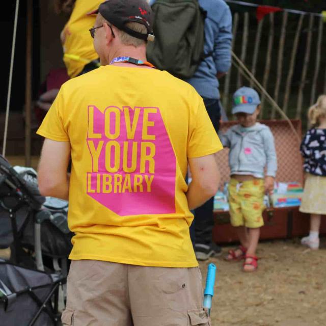 This image shows a man wearing a bright yellow T-shirt with "LOVE YOUR LIBRARY" printed in bold pink text on the back. He is standing in an outdoor setting with a crowd of people, including children and adults, around him. Some individuals are browsing books displayed in an open suitcase