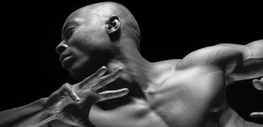 This black-and-white image shows a muscular man in a dynamic pose, with his head turned and arm raised. The lighting highlights his strong features and defined muscles against a dark background.